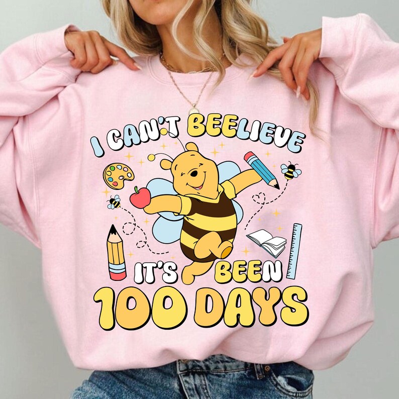 May include: Pink sweatshirt with a graphic featuring Winnie the Pooh as a bee, holding a pencil and apple. Text reads "I CAN'T BEELIEVE IT'S BEEN 100 DAYS" with school-themed illustrations. A fun, colorful design.