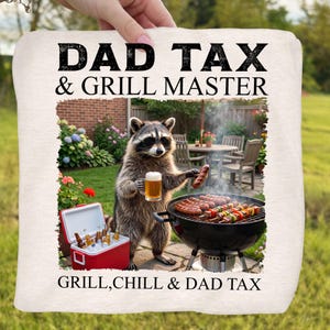 May include: A white pillow with the text "DAD TAX & GRILL MASTER" and "GRILL, CHILL & DAD TAX". The image features a raccoon wearing sunglasses, holding a beer and a sausage, grilling food.