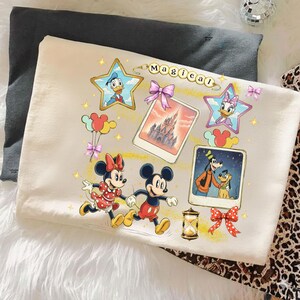 May include: Cream-colored t-shirt featuring Disney characters Mickey and Minnie Mouse, running. The shirt has a "Magical" design with Donald Duck, Daisy Duck, and Goofy. Includes a castle image and Mickey Mouse balloons.