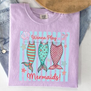 May include: A light purple Comfort Colors t-shirt with a graphic of three mermaid tails hanging on a clothesline. The tails are red and orange striped, teal with stars, and pink with scales. The text "Wanna Play Mermaids!" is above and below the tails.