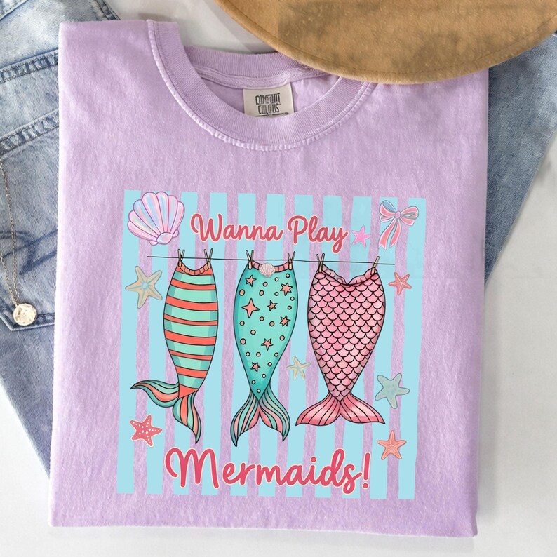 May include: A light purple Comfort Colors t-shirt with a graphic of three mermaid tails hanging on a clothesline. The tails are red and orange striped, teal with stars, and pink with scales. The text "Wanna Play Mermaids!" is above and below the tails.