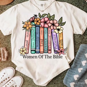 Könnte beinhalten: Weißes T-Shirt mit einem farbenfrohen Grafikdesign aus Büchern und Blumen. Die Bücher sind mit Wörtern wie "Loyal" und "Patience" beschriftet. Der Text "Women Of The Bible" steht darunter.