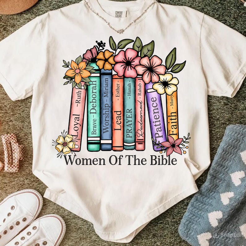 May include: White t-shirt featuring a colorful graphic of books with floral accents. The books are labeled with names like "Loyal," "Brave," and "Patience," with the text "Women Of The Bible" below.