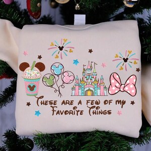 May include: Cream-colored sweatshirt with embroidered Disney-themed graphics. Features a milkshake, balloons, a castle, and a bow. Text reads "These are a few of my favorite things."