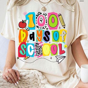 May include: Cream-colored t-shirt with a colorful graphic that reads "100 Days of School." The design includes a red apple, pencil, and paper airplane. The text is in various bright colors and fonts.