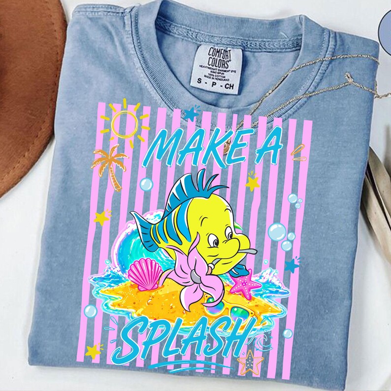 May include: A light blue t-shirt with a graphic featuring a yellow fish, the words "MAKE A SPLASH", and other sea-themed elements. The design includes pink stripes, a palm tree, and bubbles.