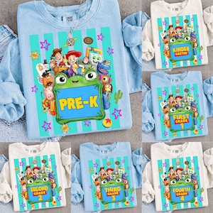May include: A light blue t-shirt featuring a graphic of Toy Story characters surrounding a green frog-shaped frame with the text PRE-K inside. The design includes vertical teal stripes, pink stars, and yellow accents on a soft cotton fabric.