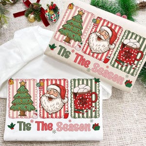 Puede incluir: Dos sudaderas blancas con un dise&ntilde;o navide&ntilde;o. Una sudadera presenta un &aacute;rbol de Navidad, Pap&aacute; Noel y una taza de chocolate caliente con el texto "Tis The Season". La otra sudadera tiene el mismo dise&ntilde;o.