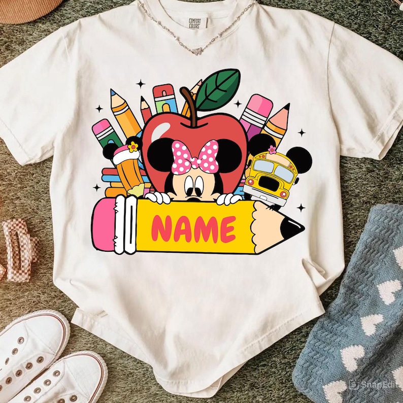 May include: White t-shirt featuring a colorful graphic of Minnie Mouse as an apple, holding a yellow pencil with the word "NAME". The design includes pencils, a school bus, and stars, perfect for back-to-school. The shirt is made of a soft material.
