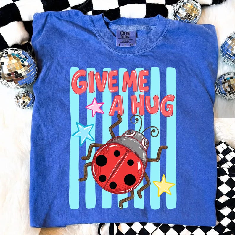 Pu&ograve; includere: Una maglietta blu con la scritta "GIVE ME A HUG" in lettere rosse. La maglietta presenta una coccinella dei cartoni animati con corpo rosso e pois neri, su uno sfondo di strisce blu e grafiche a stella. &Egrave; una maglietta del marchio Comfort Colors.
