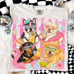 May include: White Comfort Colors t-shirt featuring a graphic of four dogs wearing sunglasses and enjoying tropical drinks and floats. The design includes pink stripes, seashells, and starfish, evoking a summer theme.