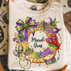 May include: Beige t-shirt featuring a colorful Mardi Gras design. The design includes a wreath with a bow, mask, maracas, drum, and a crawfish. The words "Mardi Gras" are written in a script font within the wreath.