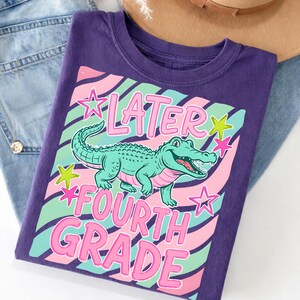 May include: A purple t-shirt with the words "Later Fourth Grade" and a cartoon alligator. The design includes pink, green, and blue stripes and stars. The shirt is folded on top of blue jeans and a brown hat.