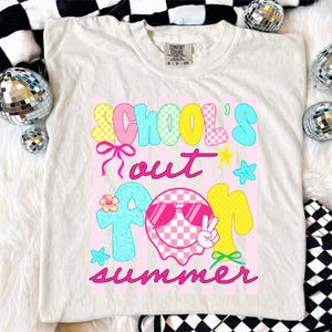 May include: Cream-colored t-shirt with a colorful graphic that reads "SCHOOL'S OUT FOR SUMMER." The design features checkered patterns, stars, and a smiley face wearing sunglasses. The shirt is a Comfort Colors brand.