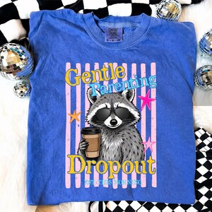Op de afbeelding: Een blauw Comfort Colors T-shirt met een wasbeer met een zonnebril en een koffiekopje. Het shirt heeft de tekst "Gentle Parenting Dropout EST. THIS MORNING" in een kleurrijk ontwerp met roze en gele accenten.