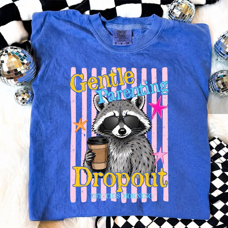 Op de afbeelding: Een blauw Comfort Colors T-shirt met een wasbeer met een zonnebril en een koffiekopje. Het shirt heeft de tekst "Gentle Parenting Dropout EST. THIS MORNING" in een kleurrijk ontwerp met roze en gele accenten.
