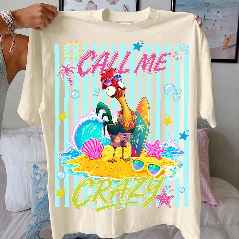 May include: Cream-colored t-shirt with a colorful graphic of a rooster wearing sunglasses and holding a surfboard. The text "CALL ME CRAZY" is displayed above and below the rooster. The design includes a beach scene with a wave, starfish, and sunglasses.