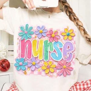 May include: Cream-colored t-shirt featuring the word "nurse" in colorful, bubble letters, surrounded by pastel-colored flower graphics. The background has a pink and white checkered pattern. A cheerful and vibrant design.