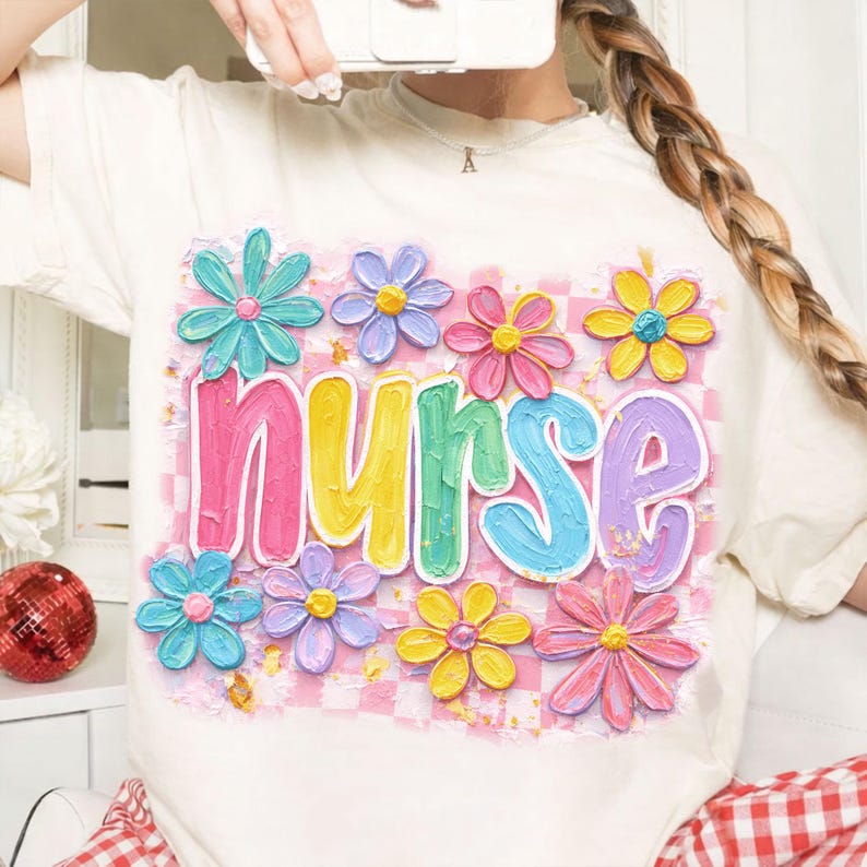May include: Cream-colored t-shirt featuring the word "nurse" in colorful, bubble letters, surrounded by pastel-colored flower graphics. The background has a pink and white checkered pattern. A cheerful and vibrant design.