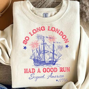 May include: Cream-colored sweatshirt with a graphic featuring a blue ship, fireworks, and text that reads "So Long London, Had a Good Run, Signed, America, *1776". The design includes red and blue accents.