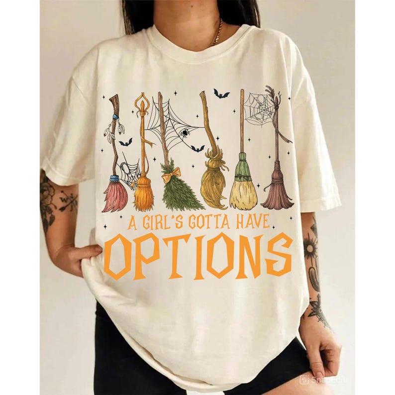 May include: Cream-colored t-shirt featuring a design of several witch brooms in various colors, with spiderwebs and bats. The text "A GIRL'S GOTTA HAVE. OPTIONS" is printed in orange. A casual, Halloween-themed tee.