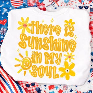 May include: White sweatshirt with the text "There is Sunshine in My Soul" in yellow, along with yellow flower and sun graphics. The sweatshirt is surrounded by a patriotic-themed background.