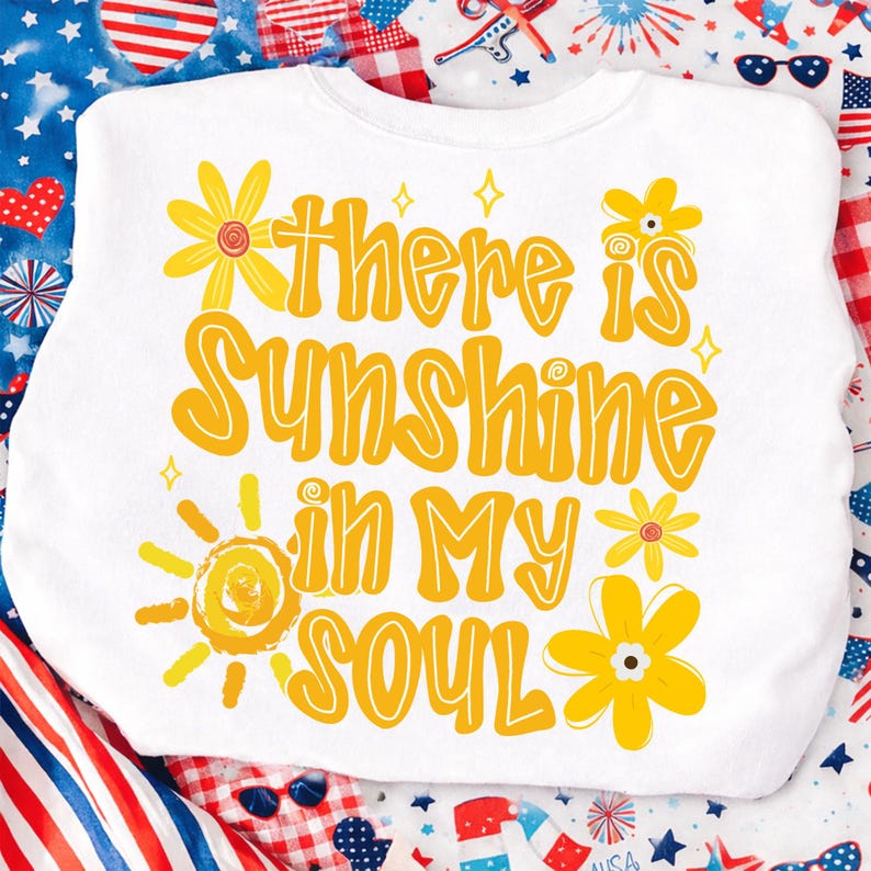 May include: White sweatshirt with the text "There is Sunshine in My Soul" in yellow, along with yellow flower and sun graphics. The sweatshirt is surrounded by a patriotic-themed background.