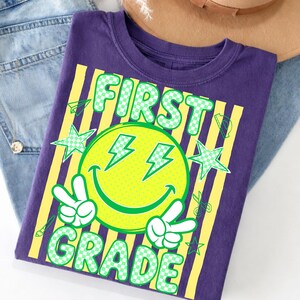 May include: A purple t-shirt with a graphic design for first grade. The design includes a yellow smiley face with lightning bolt eyes, peace signs, and the words "FIRST GRADE" in green. Yellow stripes and green stars complete the design.