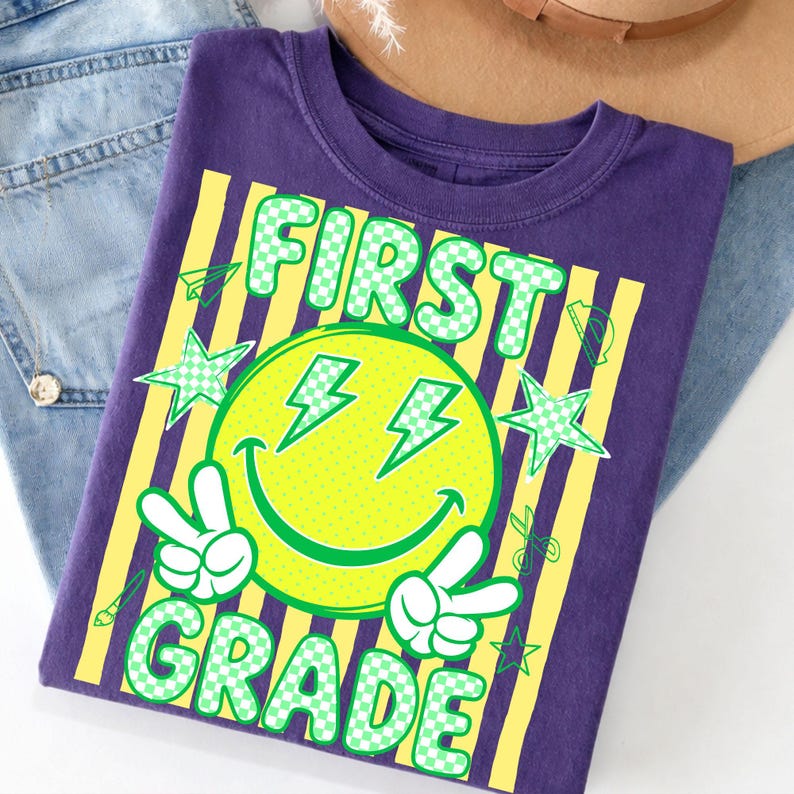 May include: A purple t-shirt with a graphic design for first grade. The design includes a yellow smiley face with lightning bolt eyes, peace signs, and the words "FIRST GRADE" in green. Yellow stripes and green stars complete the design.