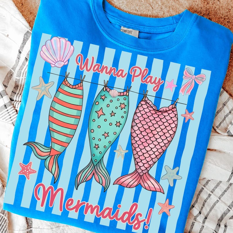 May include: A blue sweatshirt with a graphic design featuring three mermaid tails hanging on a clothesline. The text "Wanna Play Mermaids!" is displayed in pink script, with starfish and seashell accents.