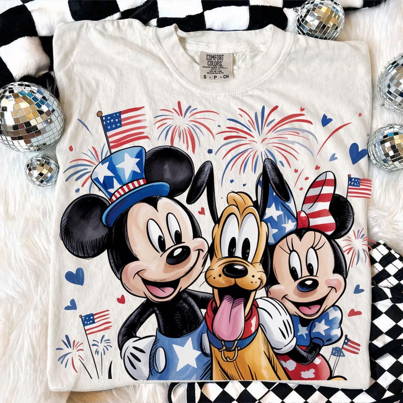 May include: Cream-colored t-shirt featuring Mickey Mouse, Minnie Mouse, and Pluto with patriotic designs. Mickey wears a blue top hat, Minnie a bow, and Pluto has a red collar. Fireworks and American flags complete the festive design.