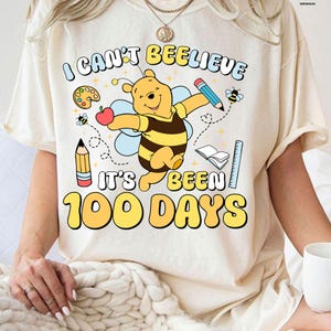 May include: Cream-colored t-shirt with the text "I can't bee-lieve it's been 100 days" and a cartoon Winnie the Pooh character dressed as a bee. The design includes pencils, an apple, a ruler, and a book.