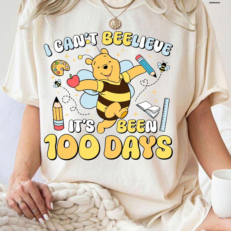 May include: Cream-colored t-shirt with the text "I can't bee-lieve it's been 100 days" and a cartoon Winnie the Pooh character dressed as a bee. The design includes pencils, an apple, a ruler, and a book.