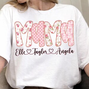 May include: White t-shirt featuring the word "Mama" in a decorative font with floral, gingham, and bow patterns. Below the word "Mama" are the names "Elle", "Taylor", and "Angela" with heart symbols.