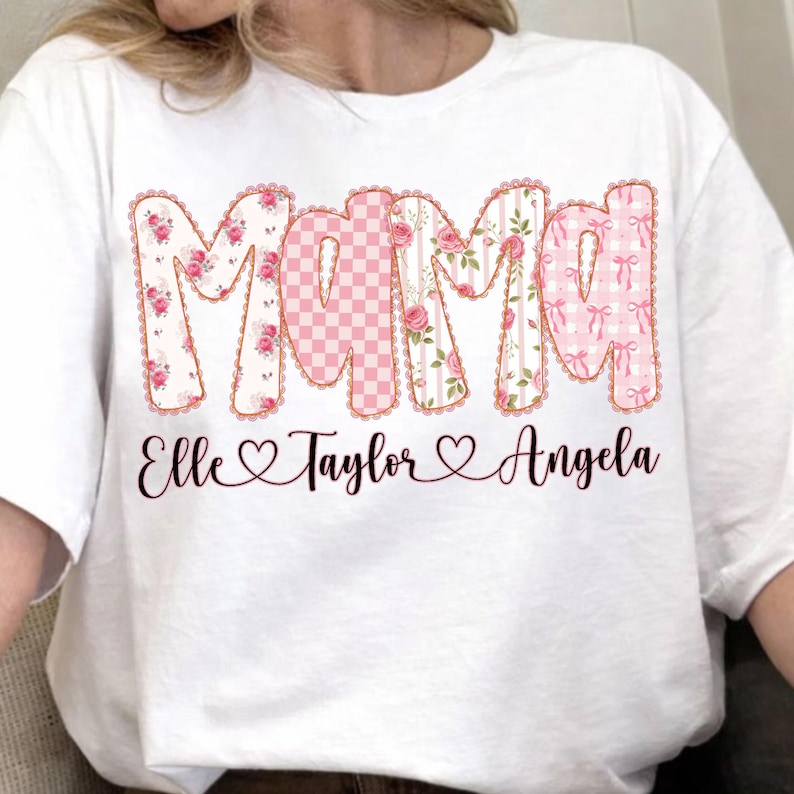 May include: White t-shirt featuring the word "Mama" in a decorative font with floral, gingham, and bow patterns. Below the word "Mama" are the names "Elle", "Taylor", and "Angela" with heart symbols.