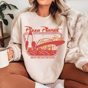 Puede incluir: Sudadera color crema con un gr&aacute;fico de Pizza Planet en rojo y amarillo. El dise&ntilde;o incluye un cohete, un restaurante y el texto "Pizza Planet" y "Serving Your Local Star Cluster".