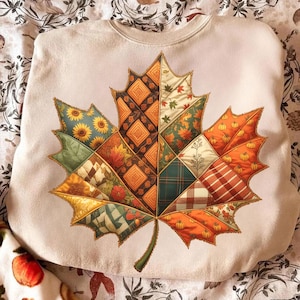 Könnte beinhalten: Cremefarbenes Sweatshirt mit einem großen Blattdesign im Patchwork-Stil. Das Blatt besteht aus verschiedenen Stoffmustern in herbstlichen Farben, darunter Orange, Grün, Gelb und Braun. Das Design umfasst Sonnenblumen, Kürbisse und Karomuster.