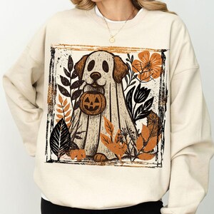 Pu&ograve; includere: Una felpa color crema con una grafica a tema Halloween. Il design presenta un cane vestito da fantasma che tiene in mano una zucca, circondato da foglie autunnali arancioni, nere e marroni. La grafica &egrave; incorniciata da un bordo nero.