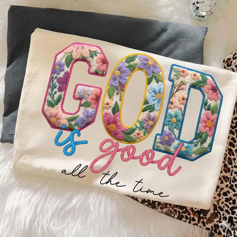 Op de afbeelding: Cr&egrave;mekleurig T-shirt met de geborduurde tekst "GOD is good all the time." De letters zijn gevuld met kleurrijke bloemmotieven in roze, paars, blauw en geel. Het shirt ligt op een witte, zachte ondergrond.