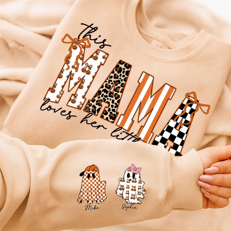 May include: Beige sweatshirt with the text "this MAMA loves her littles". The word "MAMA" is designed with pumpkin, leopard print, and striped patterns. Two ghost illustrations are on the sleeve, one with a baseball cap and the other with a bow.