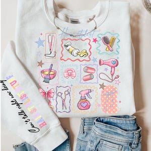 Puede incluir: Sudadera blanca con ilustraciones coloridas de herramientas de peluquería, como una plancha de pelo, un secador y unas tijeras. La manga presenta el texto "HAIRSTYLIST STUFF" y "Can't take right now doing". El diseño está rodeado de marcos coloridos.