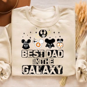 Pu&ograve; includere: Felpa color crema a tema Star Wars. Presenta disegni di personaggi ispirati a Mickey Mouse di Darth Vader, uno Stormtrooper e BB-8. Il testo recita "BEST DAD IN THE GALAXY" in un carattere invecchiato. Include una grafica della Morte Nera.