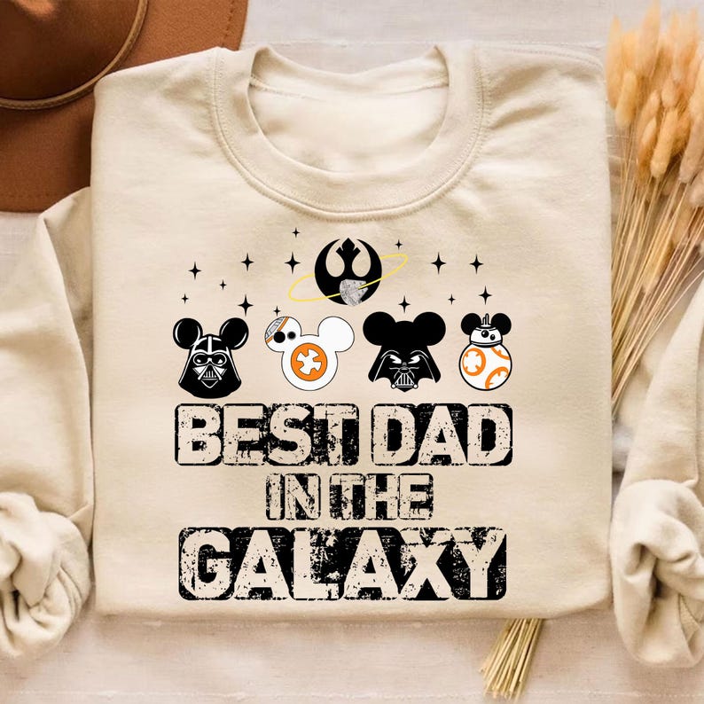Pu&ograve; includere: Felpa color crema a tema Star Wars. Presenta disegni di personaggi ispirati a Mickey Mouse di Darth Vader, uno Stormtrooper e BB-8. Il testo recita "BEST DAD IN THE GALAXY" in un carattere invecchiato. Include una grafica della Morte Nera.