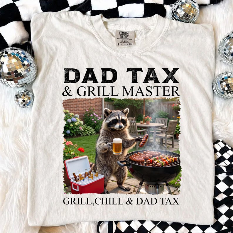 May include: Cream-colored t-shirt with the text "DAD TAX & GRILL MASTER" in black. The graphic shows a raccoon grilling with a beer and a cooler of drinks. The text "GRILL, CHILL & DAD TAX" is at the bottom.
