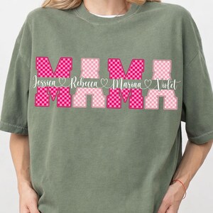 May include: Sage green t-shirt with the word "MAMA" in a pink checkered design. The names Jessica, Rebecca, Marina, and Violet are printed below the word "MAMA" in a cursive font.