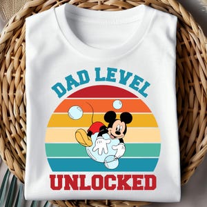 Pu&ograve; includere: T-shirt bianca con grafica di Topolino in una bolla, con la scritta "DAD LEVEL UNLOCKED". Il design presenta un tramonto retr&ograve; con strisce arancioni, gialle e turchesi. Il testo &egrave; blu e rosso.
