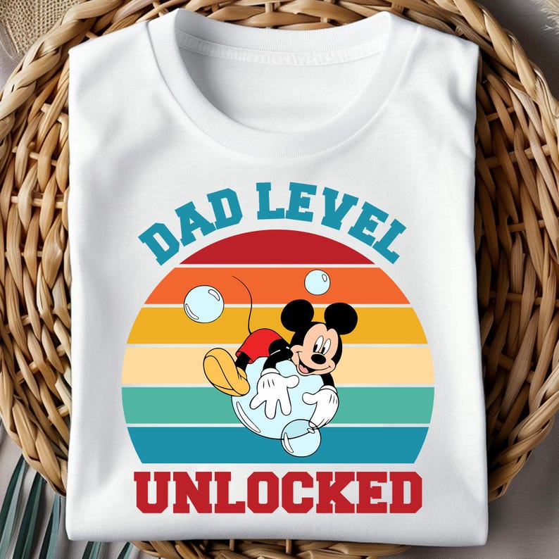 Pu&ograve; includere: T-shirt bianca con grafica di Topolino in una bolla, con la scritta "DAD LEVEL UNLOCKED". Il design presenta un tramonto retr&ograve; con strisce arancioni, gialle e turchesi. Il testo &egrave; blu e rosso.