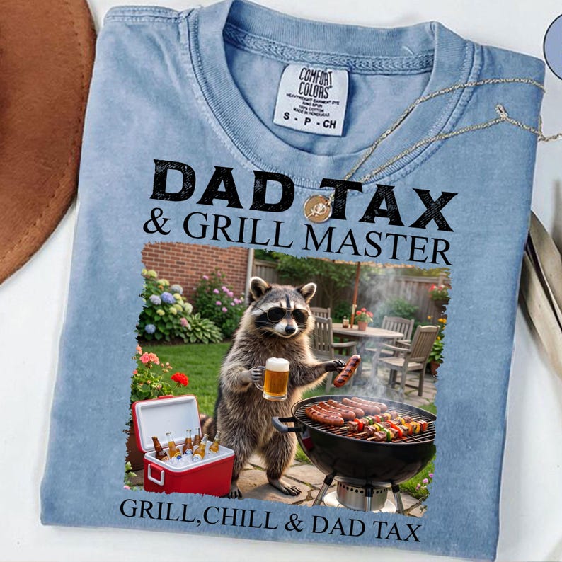 May include: Light blue Comfort Colors t-shirt with the text "DAD TAX & GRILL MASTER". The graphic shows a raccoon wearing glasses, holding a beer and a sausage, grilling. A red cooler with drinks is visible.