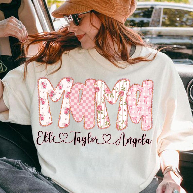 May include: Cream-colored t-shirt with the word "Mama" in pink floral and gingham patterns. The names "Elle", "Taylor", and "Angela" are printed below the word "Mama".