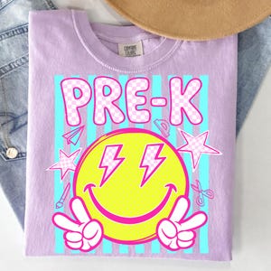 May include: A light purple t-shirt with the word "PRE-K" in pink checkered letters. The design includes a yellow smiley face with lightning bolt eyes and peace signs, along with school-themed graphics.